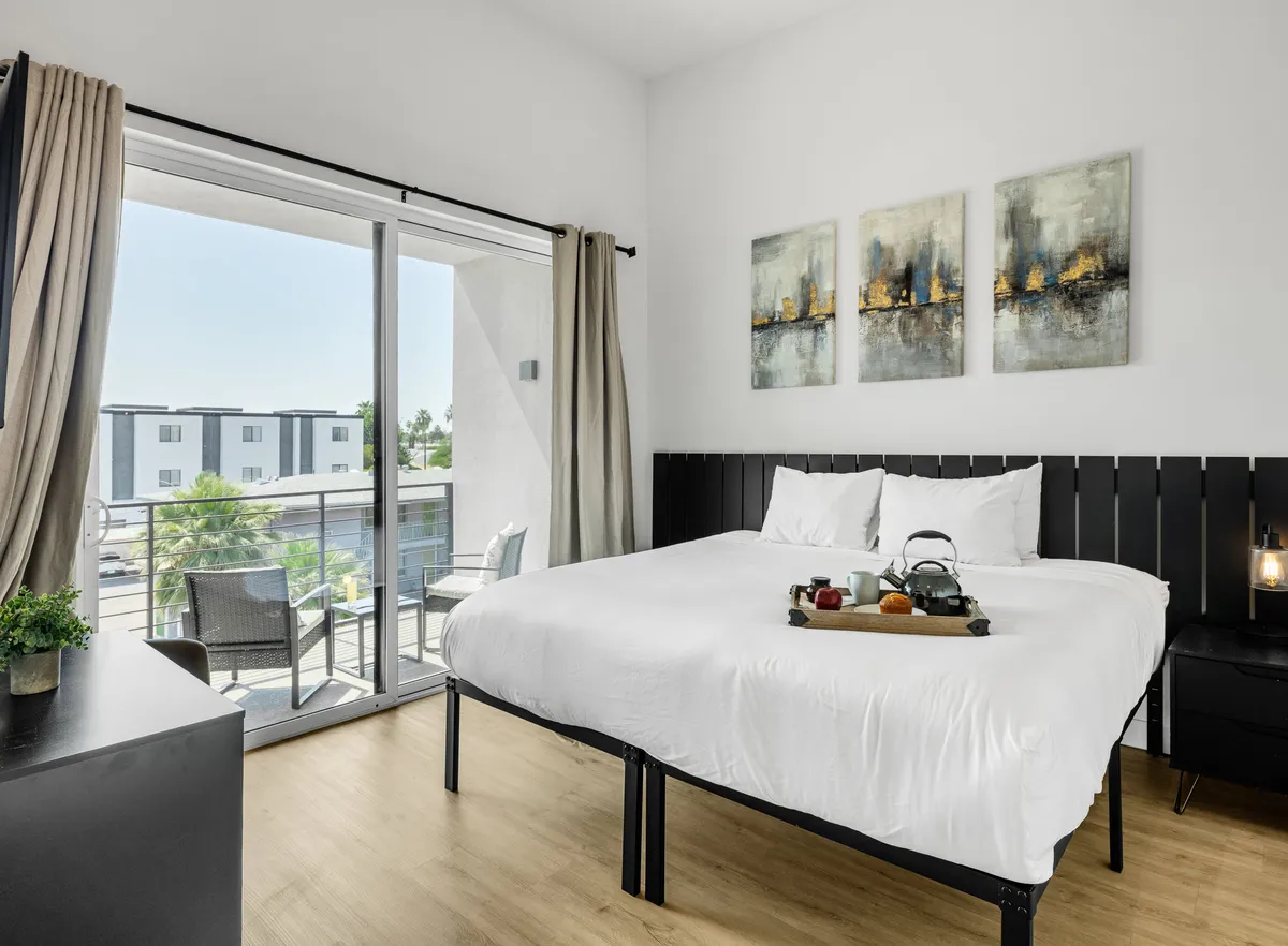 A tranquil bedroom scene with a plush bed dressed in white linens, elegant artwork above, and a balcony with cozy chairs visible through sliding doors. The room exudes comfort and a connection to outdoor relaxation.