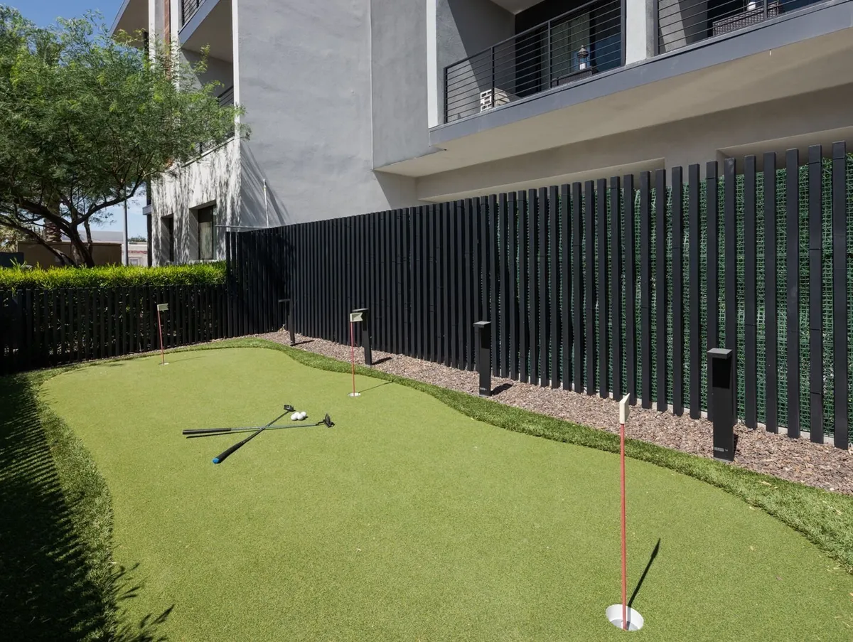 An exterior shot of a well-maintained putting green, modern architecture, landscaped grounds, and a secure fence. The area is inviting for casual golf practice or outdoor family gatherings, with sunny skies overhead.