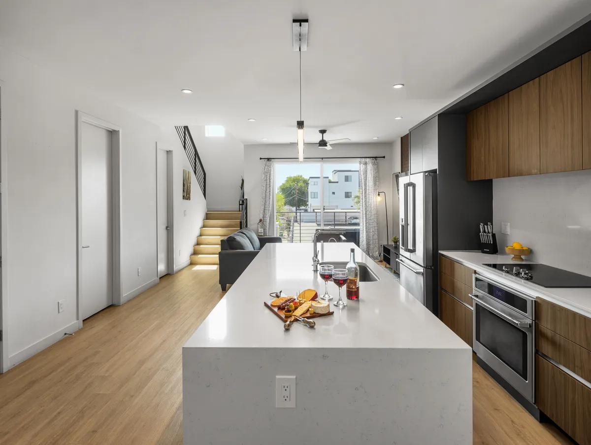 A modern, open-concept space featuring a kitchen island with stools, stainless steel appliances, and a visible living area with a plush sofa. The design blends the kitchen and living room, accented by a staircase and ample natural light from large windows.