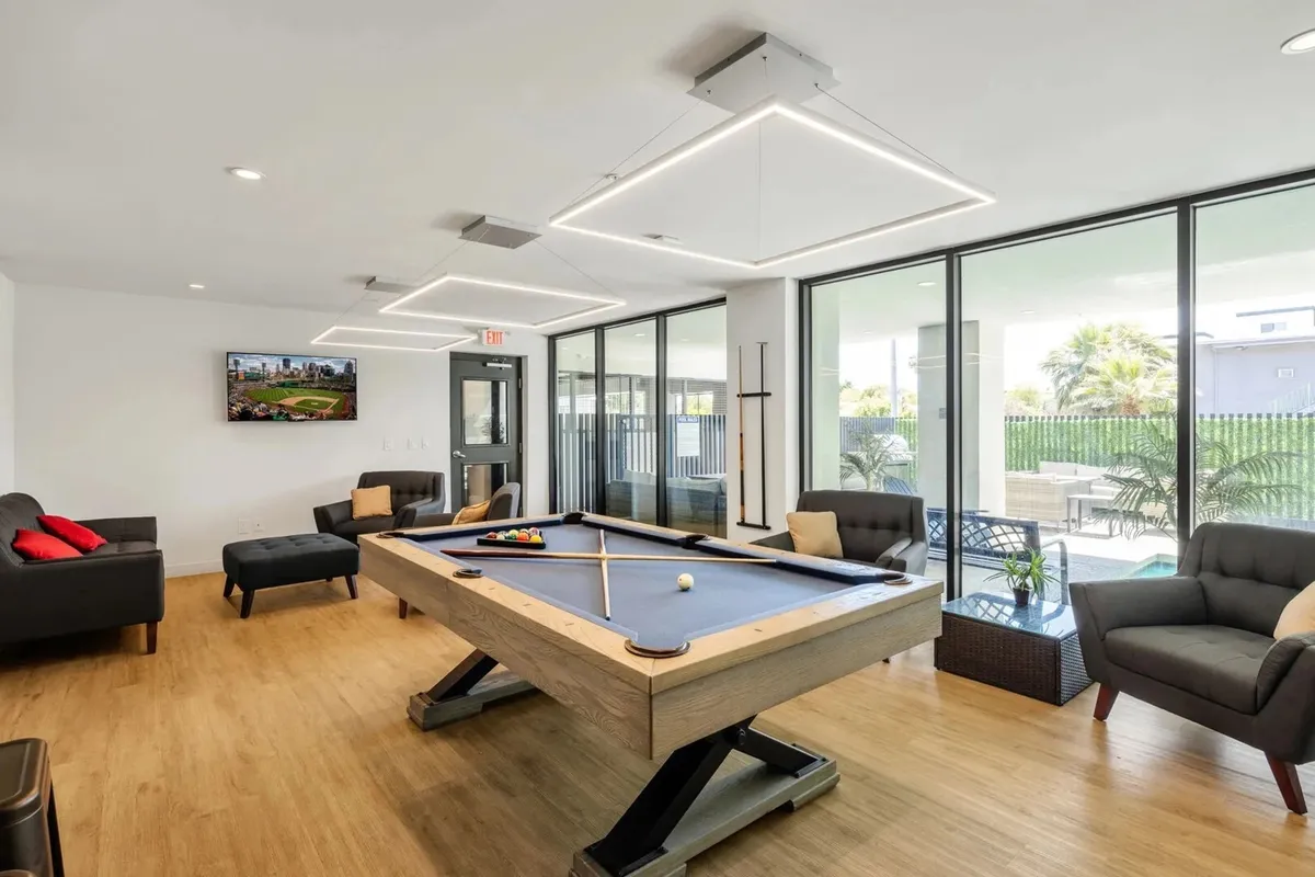 A bright, friendly clubhouse interior with a pool table, comfortable seating, large windows letting in natural light, and a television. The scene is set for both leisure and social gatherings.