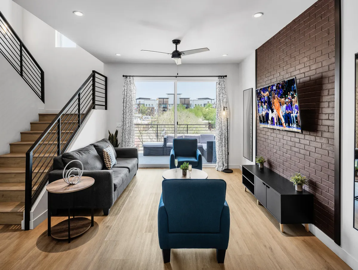 A spacious, modern living area featuring a comfortable sofa, blue accent chairs, a large TV, and a prominent wooden staircase leading to upper floors. Large windows allow for natural light, and the open floor plan hints at seamless access to other spaces within the unit.